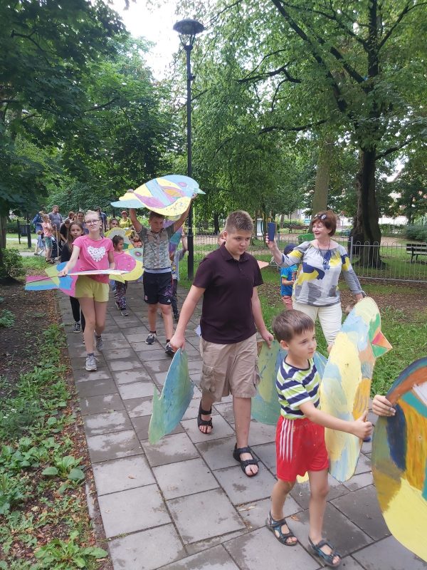 Przemarsz grupy dzieci z kolorowymi ptakami przez park