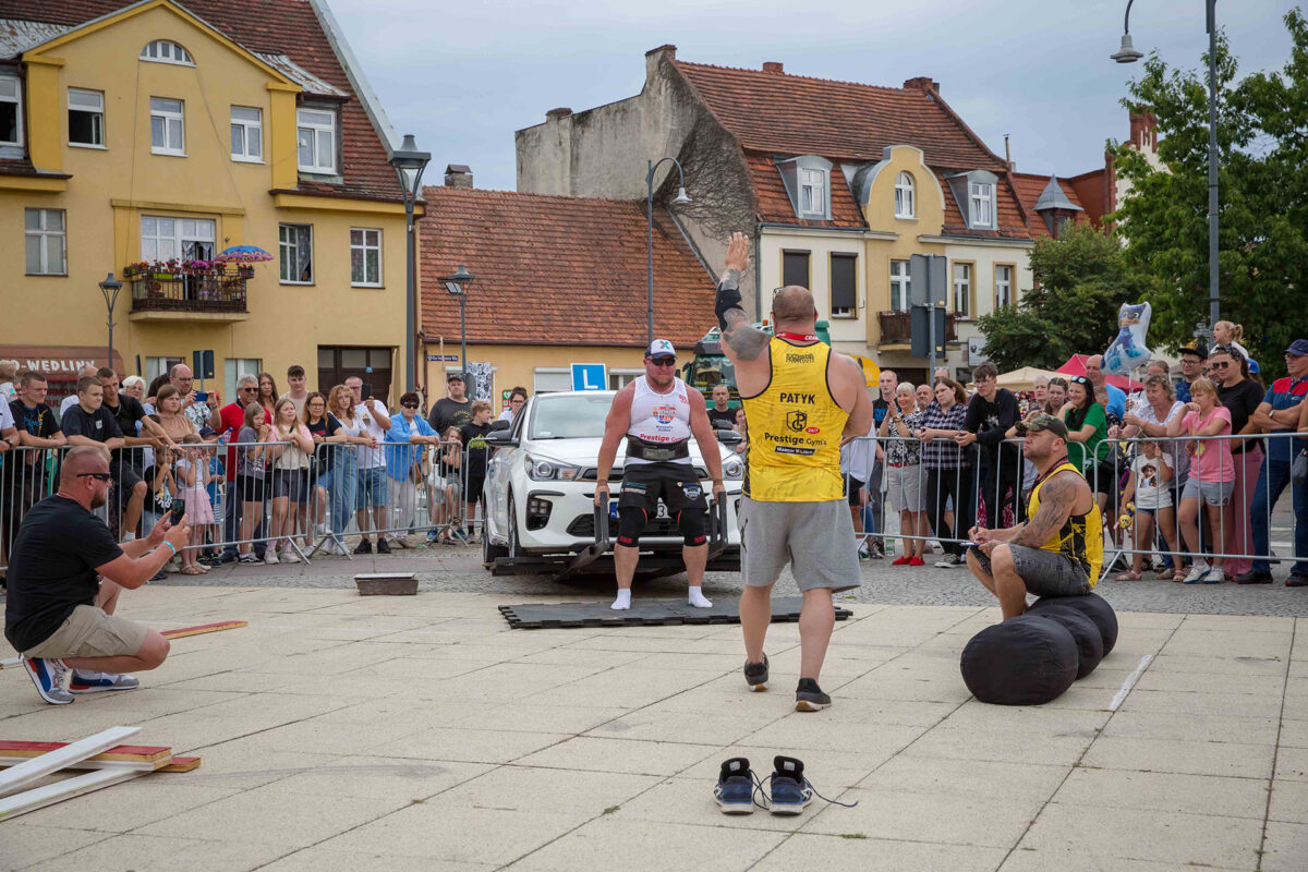 Strongman podnosi auto, publiczność w tle.