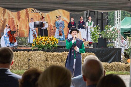Mężczyzna z zespołu ludowego przy mikrofonie, w tle scena dożynkowa z muzykami z kapeli ludowej.
