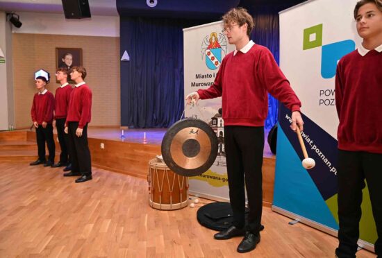 Chórzysta grający na instrumencie perkusyjnym, w tle inni chórzyści.