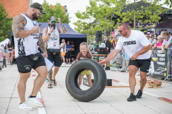 Dziewczynka przerzuca oponę w towarzystwie strongmanów.