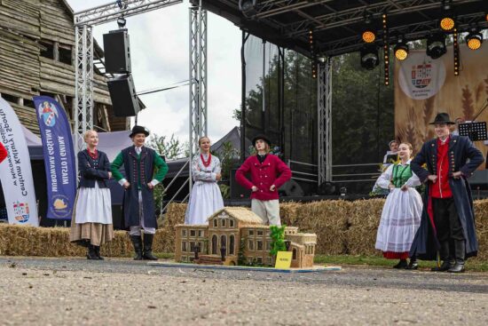 Grupa osób z zespołu ludowego stoi przy wieńcu dożynkowym.