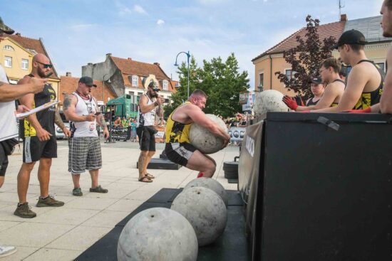 Strongman podnosi ciężar w towarzystwie innych strongmanów.
