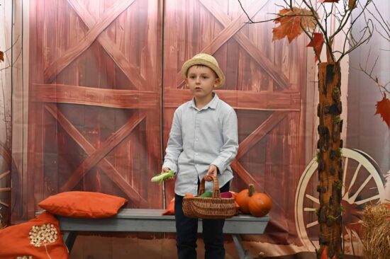 Chłopiec w kapeluszu , trzymający koszyk z warzywami  recytuję wiersz na tle jesiennej scenografii