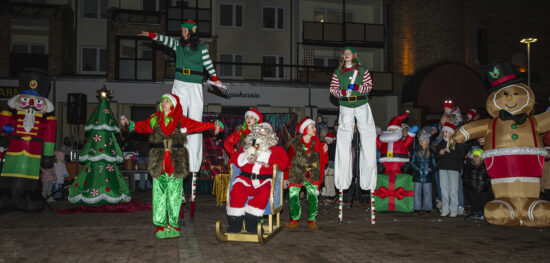 Na pierwszym planie szczudlarze, elfy i Mikołaj siedzący na saniach na tle świątecznej scenografii, w tle publiczność