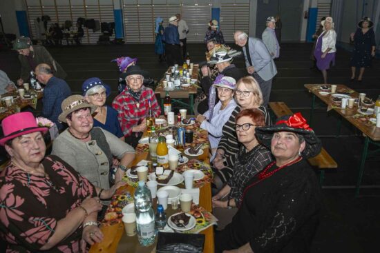 Seniorzy siedzący przy stolikach w tle grupa ludzi bawiących się na sali