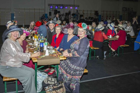 Grupa osób w kapeluszach siedzą przy stole i pozują do zdjęcia, w tle pozostali uczestnicy balu i scena