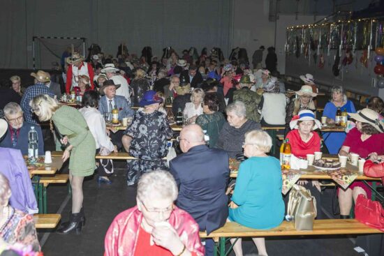 Zdjęcie sali z seniorami siedzącymi przy stolikach, w tle kolorowa scenografia