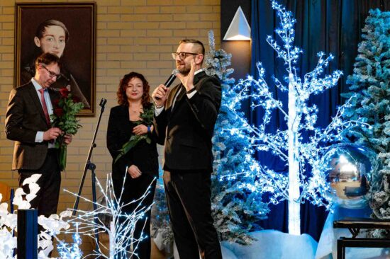 mężczyzna z mikrofonem na scenie, obok kobieta i mężczyzna z kwiatami