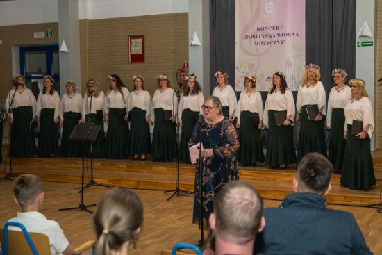 W tle Żeński Chór Canzona w białych bluzkach i zielonych spódnicach. Przed nimi przy mikrofonie stoi dyrygent Elżbieta Wtorkowska