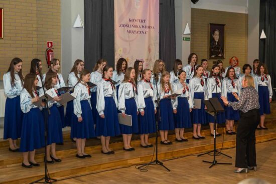 Chór dziewczęcy Canzona -grupa starsza. Dziewczynki ubrane są w błękitne bluzki i niebieskie spódnice. Przed nimi dyrygent Adrianna Wtorkowska – Kubińska