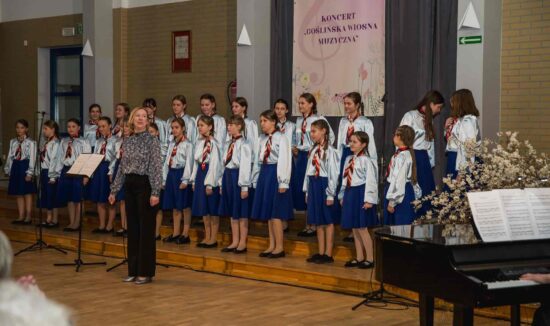 Chór dziewczęcy Canzona -grupa młodsza. Dziewczynki ubrane są w błękitne bluzki i niebieskie spódnice. Przed nimi dyrygent Adrianna Wtorkowska – Kubińska