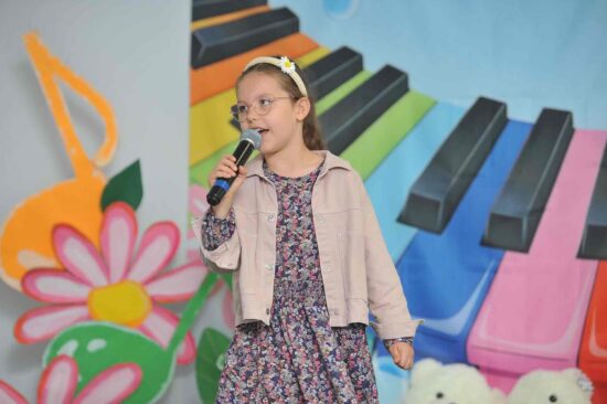 Na tle kolorowej scenografii stoi dziewczynka w sukience w kwiaty w ręku trzyma mikrofon