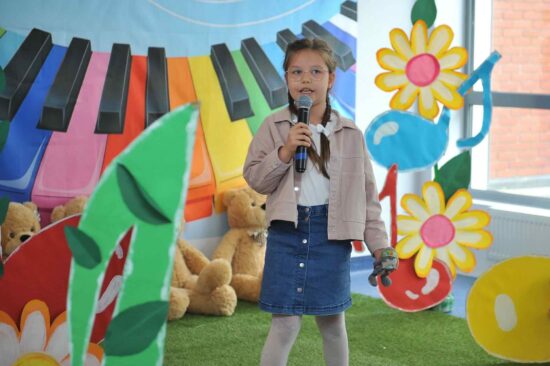 Na tle kolorowej scenografii stoi dziewczynka z warkoczykami,  w dżinsowej spódniczce i różowej kurtce z mikrofonem w ręku