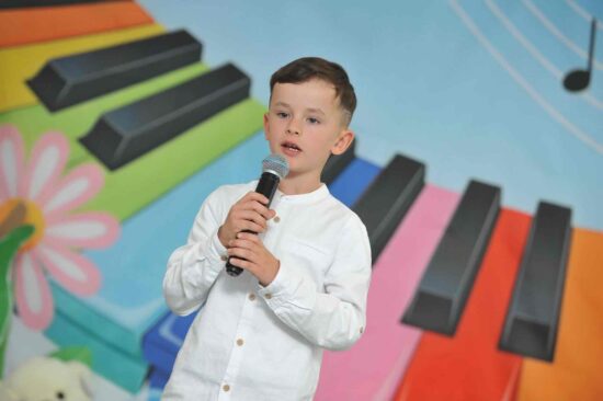 Na tle kolorowej scenografii stoi u chłopiec w białej koszuli w ręku trzyma mikrofon