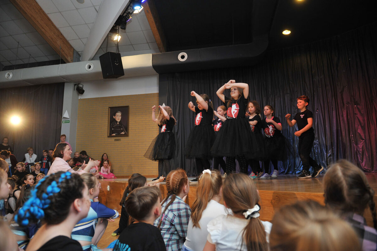 Na scenie grupa dzieci w czarnych strojach, pod sceną siedzą kibicujące dzieci