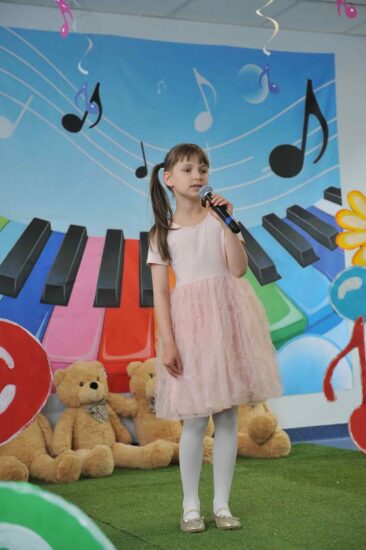 Na tle kolorowej scenografii stoi dziewczynka w różowej sukience stoi z mikrofonem w ręku