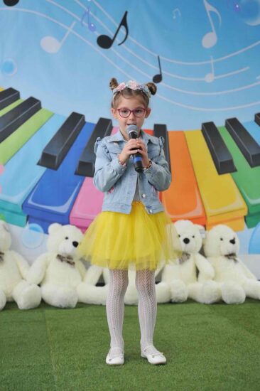 a tle kolorowej scenografii stoi dziewczynka , ma na głowie wianek, ubrana jest w żółtą spódniczkę i dżinsową kurtkę w ręku trzyma mikrofon