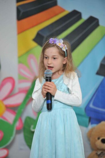 Na tle kolorowej scenografii  stoi dziewczynka w niebieskiej sukience i z wiankiem na włosach ma mikrofon w rękach