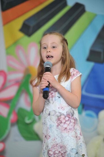 Na tle kolorowej scenografii stoi dziewczynka w sukience w kwiaty w ręku trzyma mikrofon