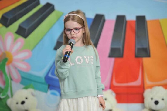Na tle kolorowej scenografii stoi dziewczynka w opasce na głowie w ręku trzyma mikrofon