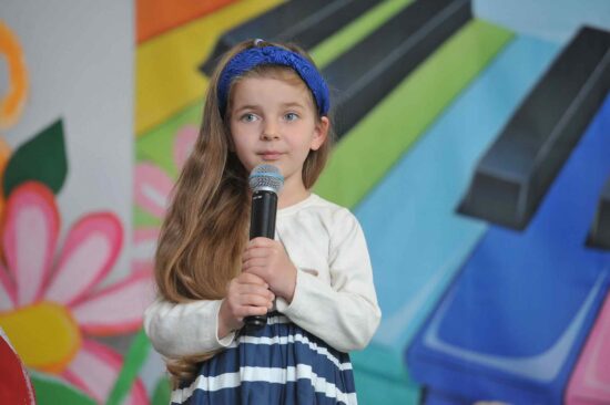 Na tle kolorowej scenografii stoi dziewczynka z długimi ,brązowym włosami  i w niebieskiej opasce na głowie  z mikrofonem w ręku