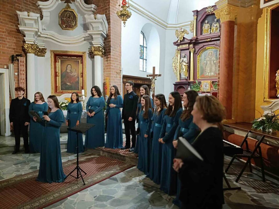 Jedna z chórzystek stoi i trzyma mikrofon, pozostali członkowie chóru w raz z dyrygentka stoją za nią