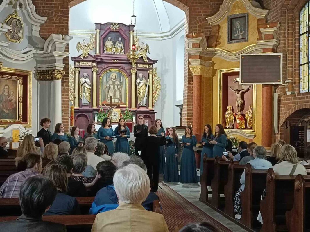 Chór stoi przed ołtarzem głównym, po bokach widać siedzącą publiczność w ławkach