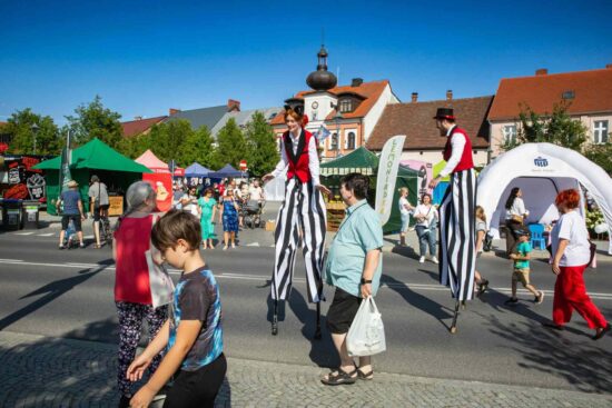 Kobieta i mężczyzna w czerwonych kamizelkach i biało czarnych spodniach na szczudłach, przechadzają się po ulicy wśród mieszkańców