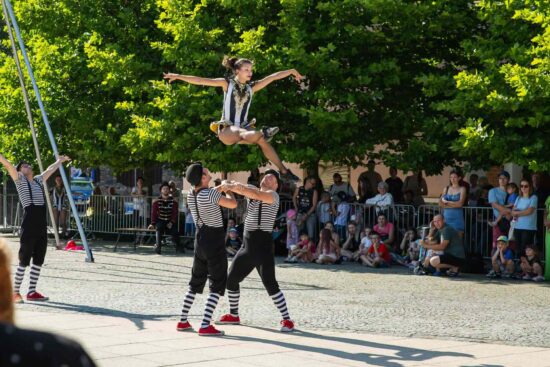 Akrobaci, dwóch mężczyzn stoi trzymając się za ręce, nad nimi kobieta - element pokazów w ramach spektaklu ulicznego Cyrkowy Street Show